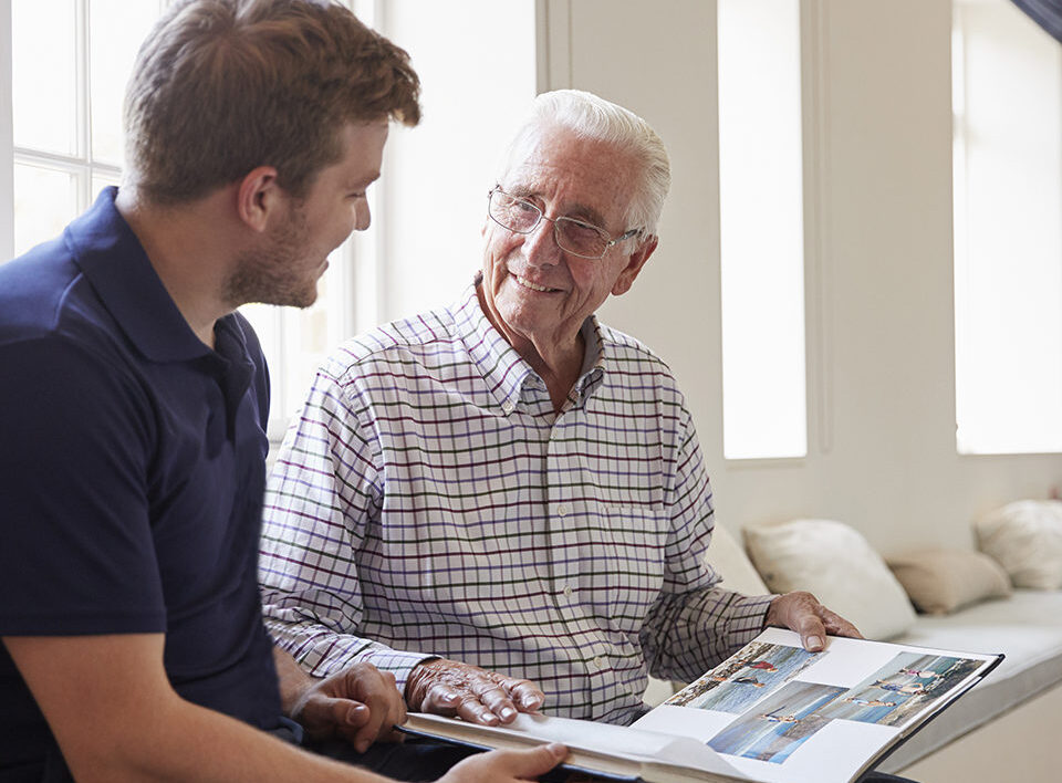 Caregiver and senior man smiling and looking at photo album together.