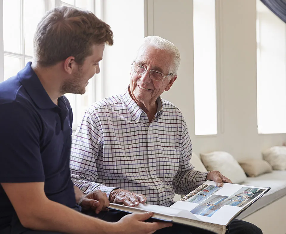 Caregiver and senior man smiling and looking at photo album together.