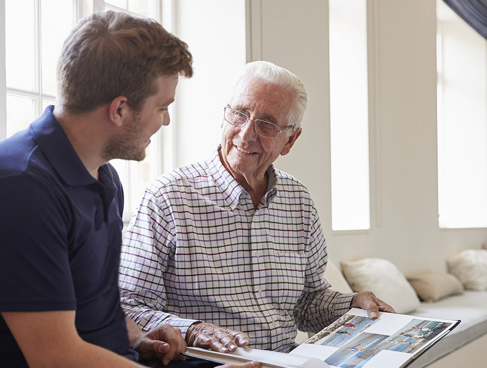 Caregiver and senior man smiling and looking at photo album together.