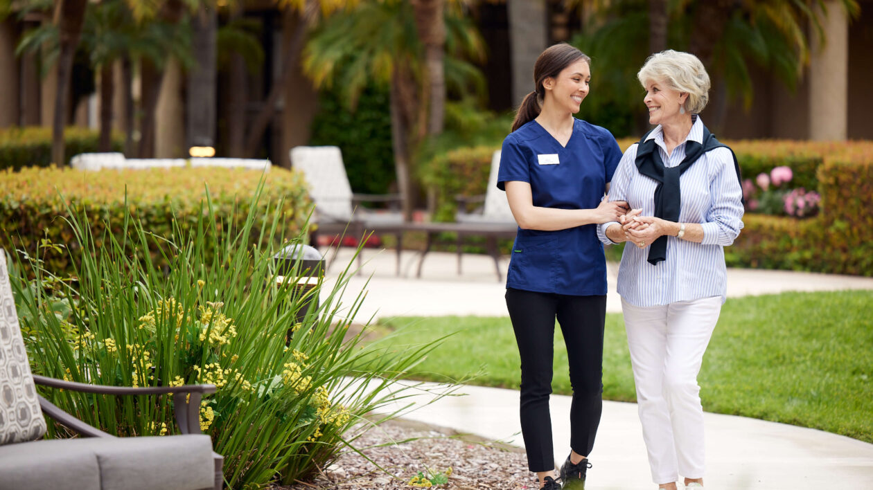 A caregiver walking in arms with an elderly woman through the courtyard.