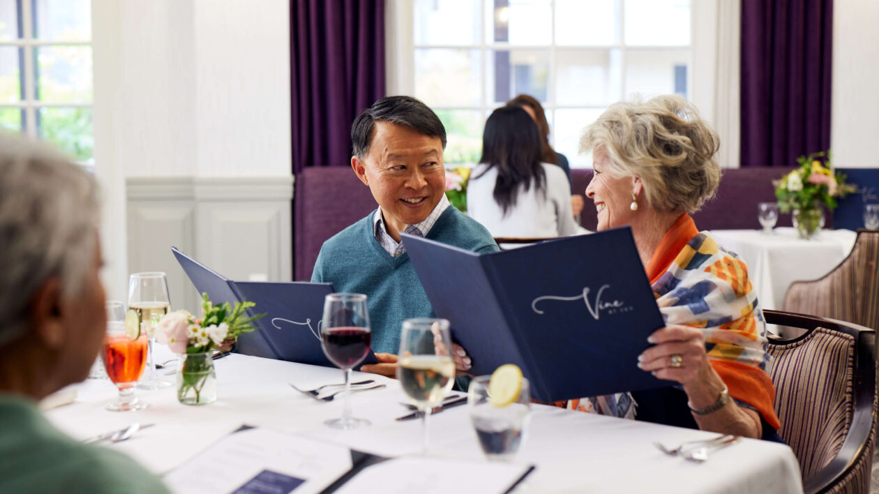 A man and woman sitting side by side at a dining table, with glasses of wine in front of them, looking at each other while trying to decide what to order from the menu.