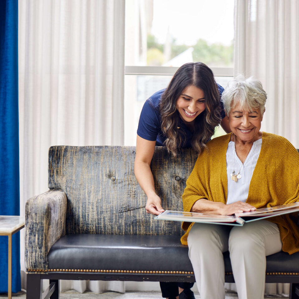 A caregiver and elderly woman looking through a large photobook together on a sofa.