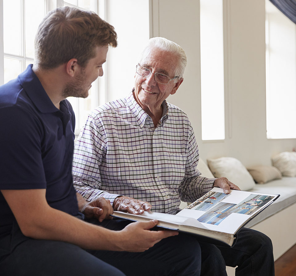 Caregiver and senior man smiling and looking at photo album together.