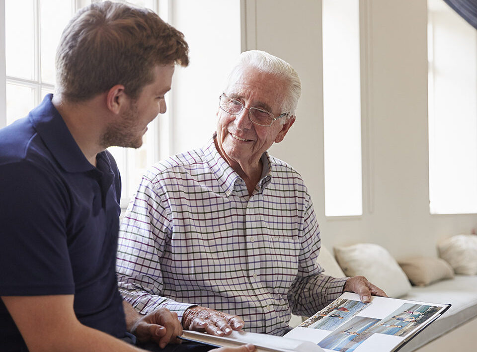 Caregiver and senior man smiling and looking at photo album together.