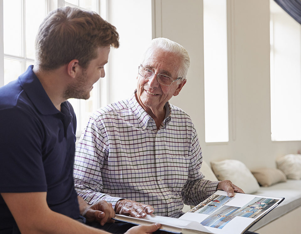 Caregiver and senior man smiling and looking at photo album together.