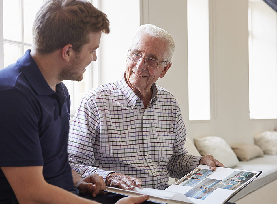 Caregiver and senior man smiling and looking at photo album together.