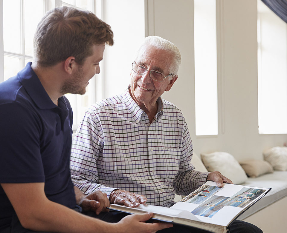Caregiver and senior man smiling and looking at photo album together.