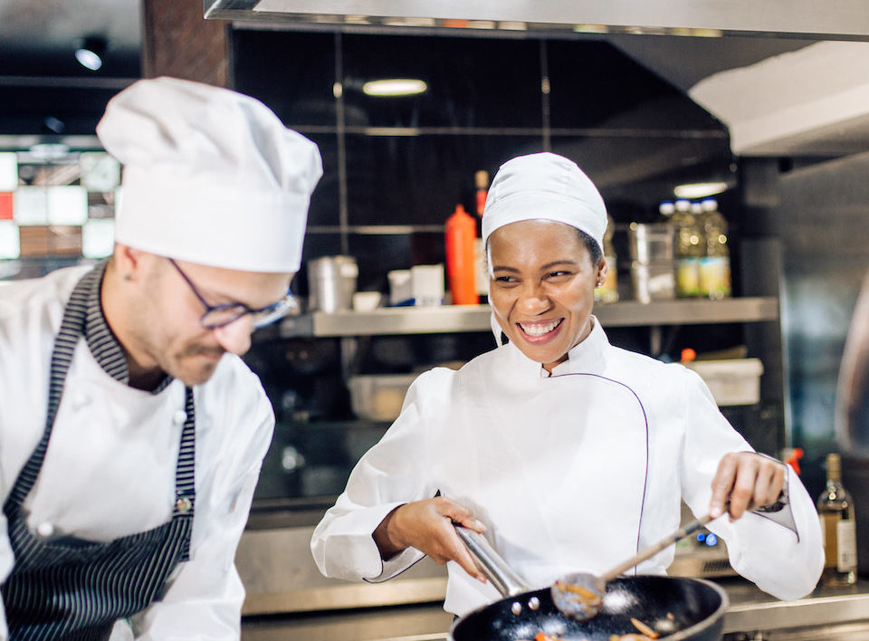 Two chefs in a kitchen, one is sauteing pasta