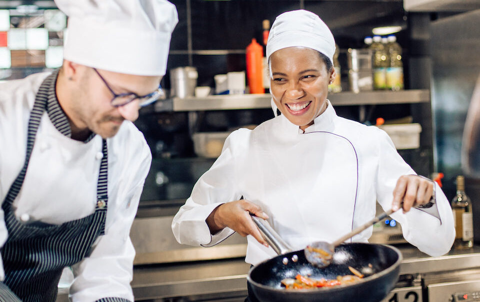 Two chefs in a kitchen, one is sauteing pasta