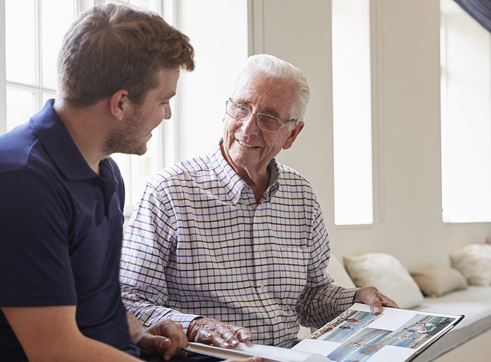 Caregiver and senior man smiling and looking at photo album together.