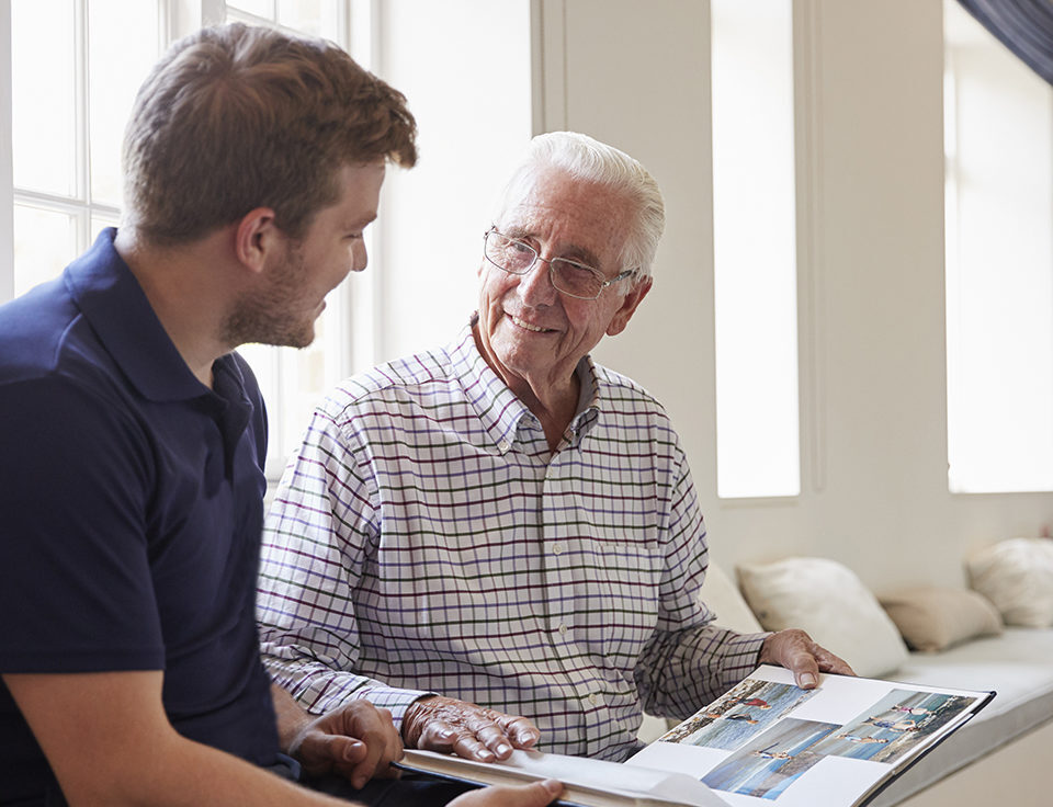 Caregiver and senior man smiling and looking at photo album together.