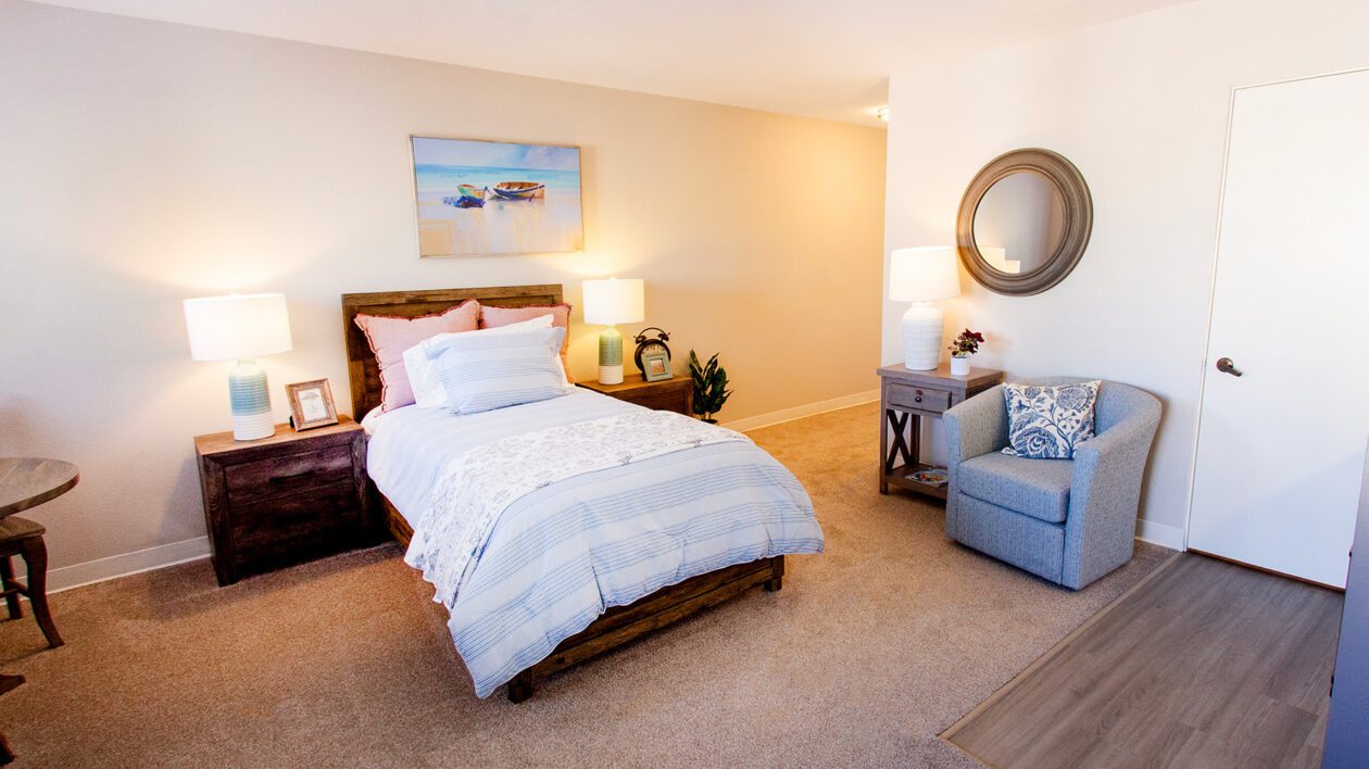 A photo of a cozy bedroom unit with a twin bed, carpet, and a lounge chair.