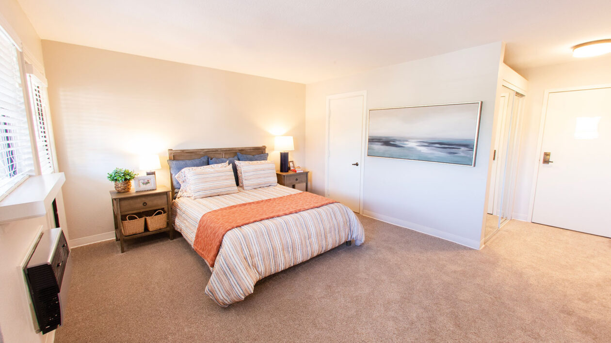 A photo of a carpeted bedroom unit with a queen bed and a painting of seawater on the wall.