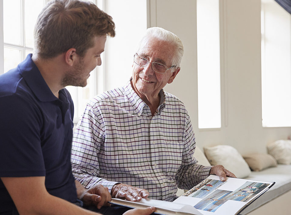 Caregiver and senior man smiling and looking at photo album together.