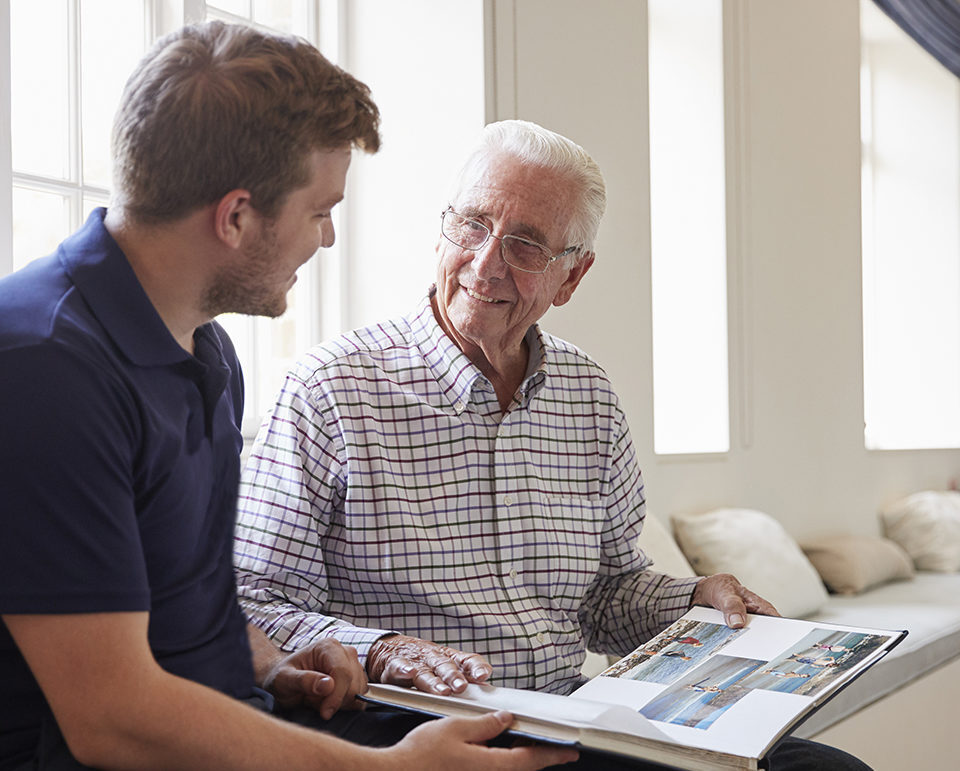 Caregiver and senior man smiling and looking at photo album together.