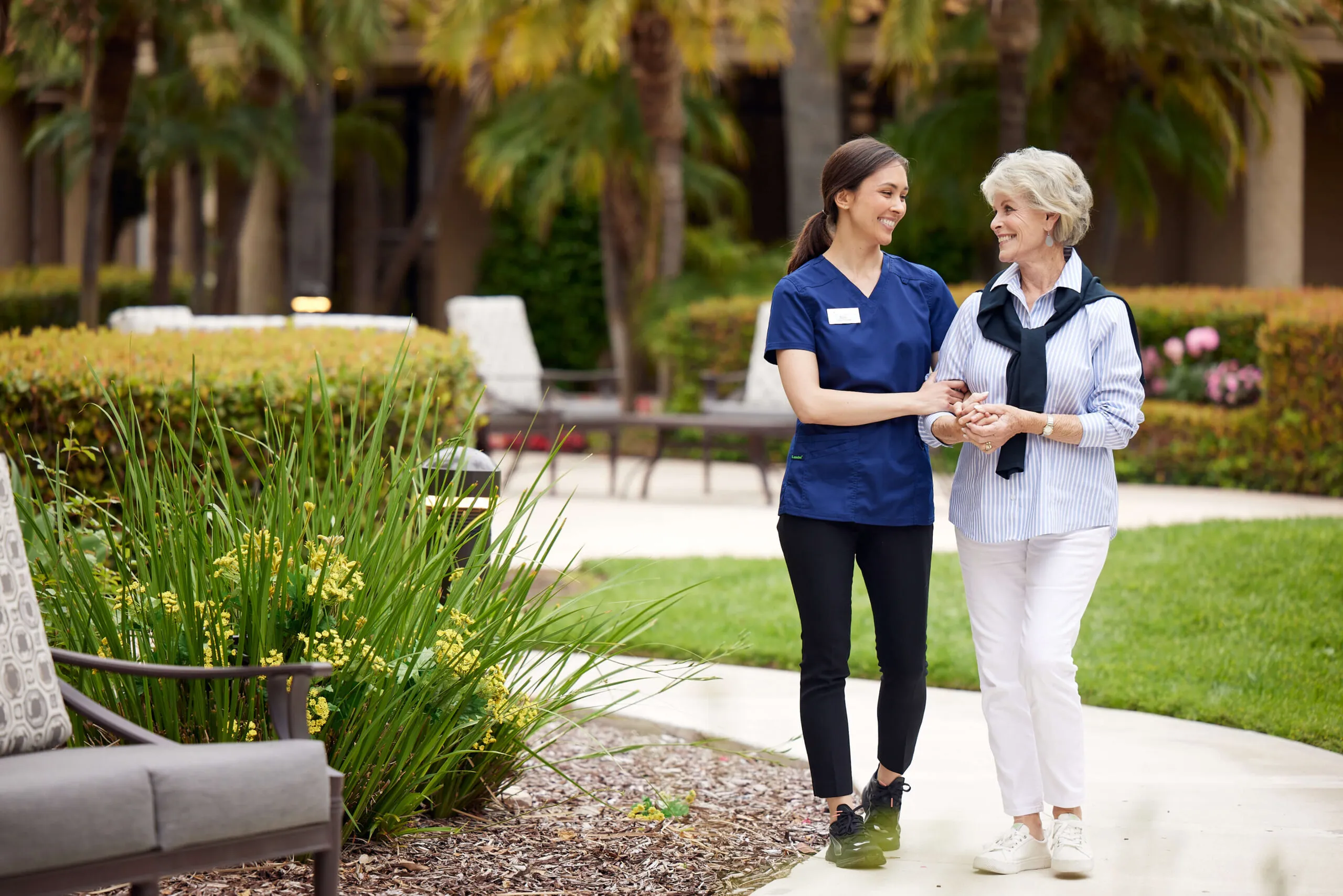 A caregiver walking in arms with an elderly woman through the courtyard.