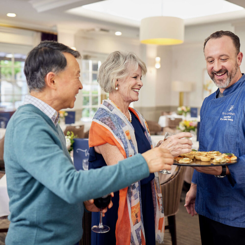 An Ivy server sharing appetizers on a platter with an elderly man and woman in the dining space.