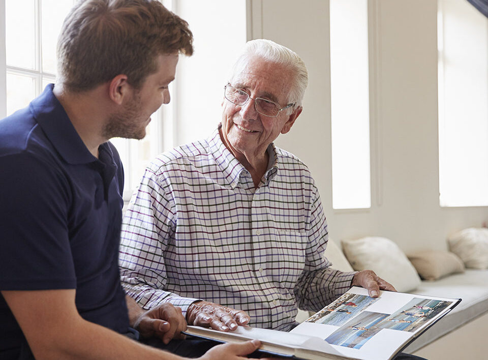 Caregiver and senior man smiling and looking at photo album together.