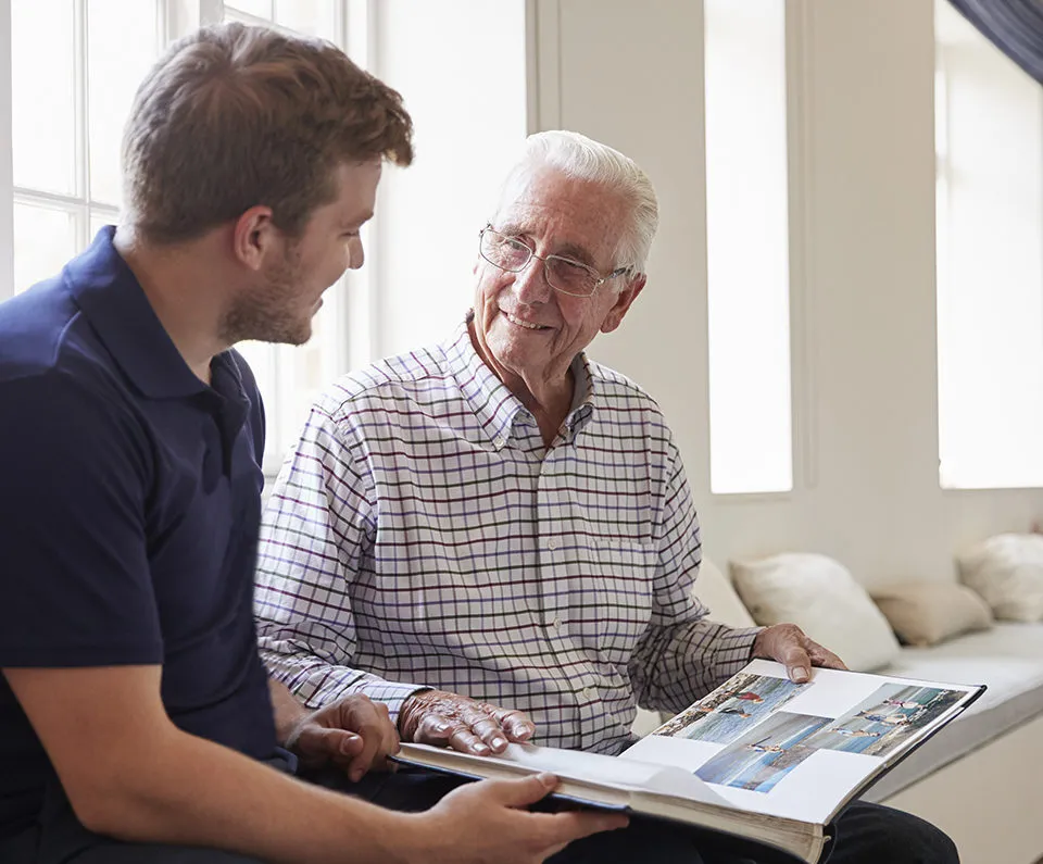 Caregiver and senior man smiling and looking at photo album together.