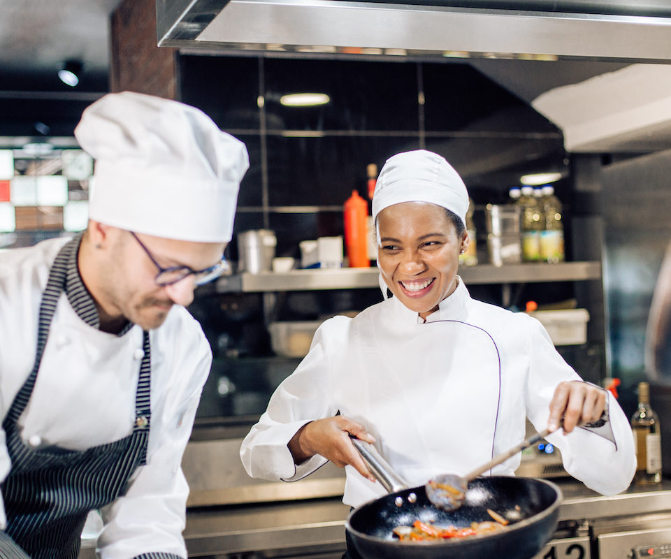 Two chefs smiling as they fry vegetables in a frying pan in the kitchen.
